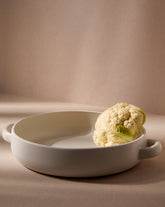A white stoneware serving plate with handles, featuring a minimalist design, displayed with a piece of cauliflower on it, set against a neutral background.