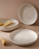 A set of three white stoneware plates, with one plate containing some pasta and cutlery, displayed against a neutral background.