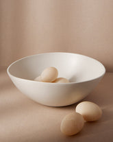 A white stoneware serving bowl with a matte finish on a neutral background, containing what appears to be a few eggs or round items, with the bowl's artisanal craftsmanship highlighted.