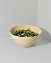A beige serving bowl with a speckled cream finish, containing salad, placed on a white surface.
