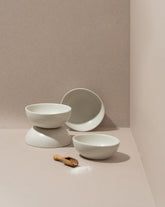 A set of white stoneware condiment bowls with a matte finish, displayed against a neutral background. One bowl is upright, while the others are stacked behind it, and there is a small wooden spoon in front.