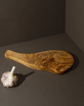 A natural olive wood cheese board with an organic shape and handle, displayed alongside a bulb of garlic.