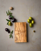A rectangular olive wood chopping board with a groove design, accompanied by various small olive fruits and a sprig of rosemary on a light background.