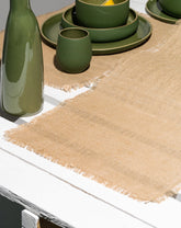 A burlap table runner laid out on a table, with green dinnerware set on top of it. The runner has a textured, natural beige color with frayed edges.