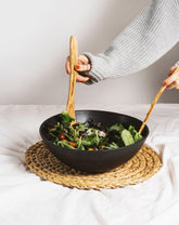 A person's hands using olive wood serving forks to serve salad from a black stoneware bowl, which is placed on a beige mat.