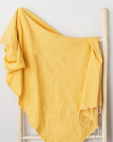 A Mustard cotton throw blanket draped over a wooden rack, with part of the blanket visible on the ground.