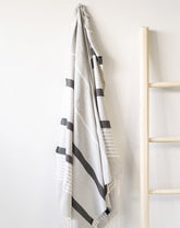 A gray fouta towel with black stripes is hanging on a wall next to a wooden ladder.