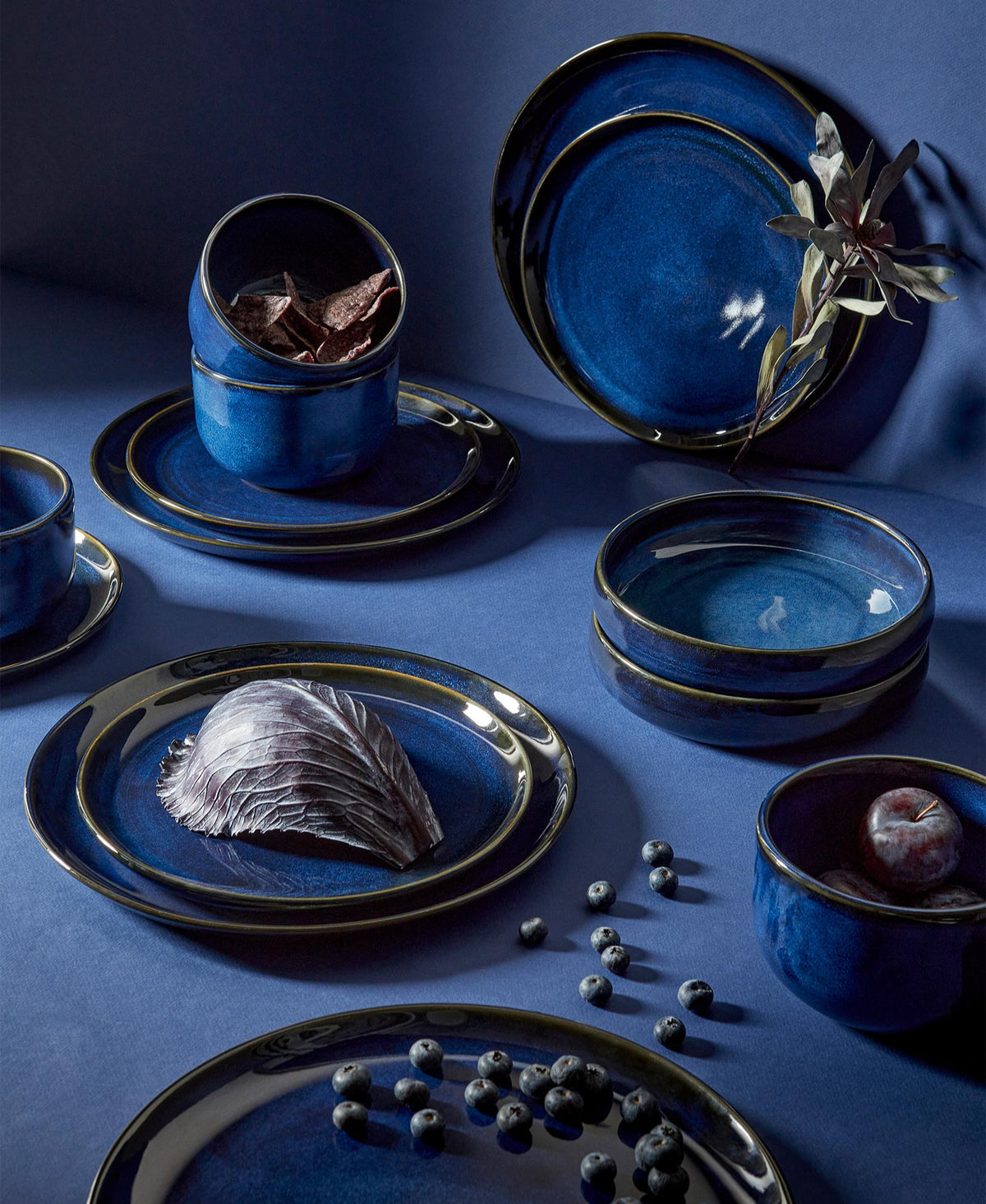 A set of beige stoneware dinnerware including flat dinner plates, flat salad plates, pasta plates/bowls, and dinner bowls, displayed with garlic and nuts on the plates.