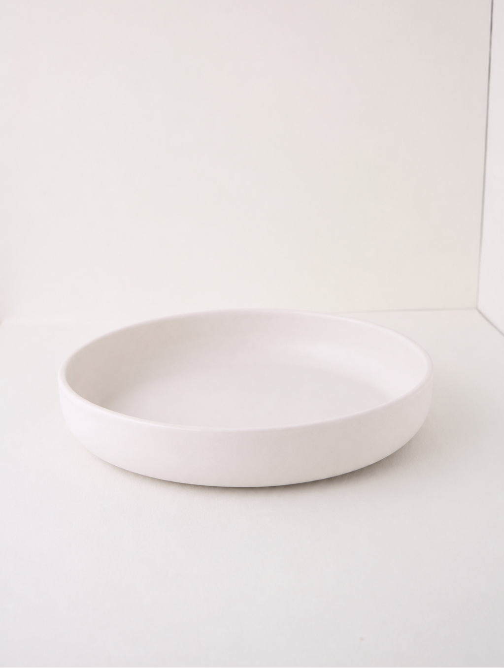 A minimalist, shallow round ceramic plate in matte white, photographed against a clean white background.