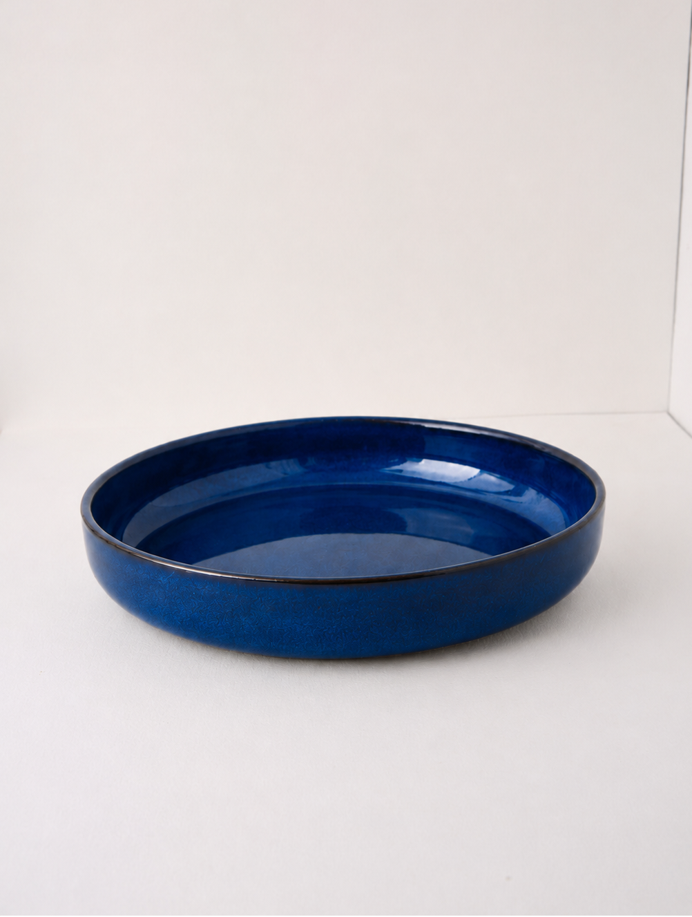 A glossy cobalt blue ceramic shallow bowl or dish, photographed from a side angle against a plain white studio background.