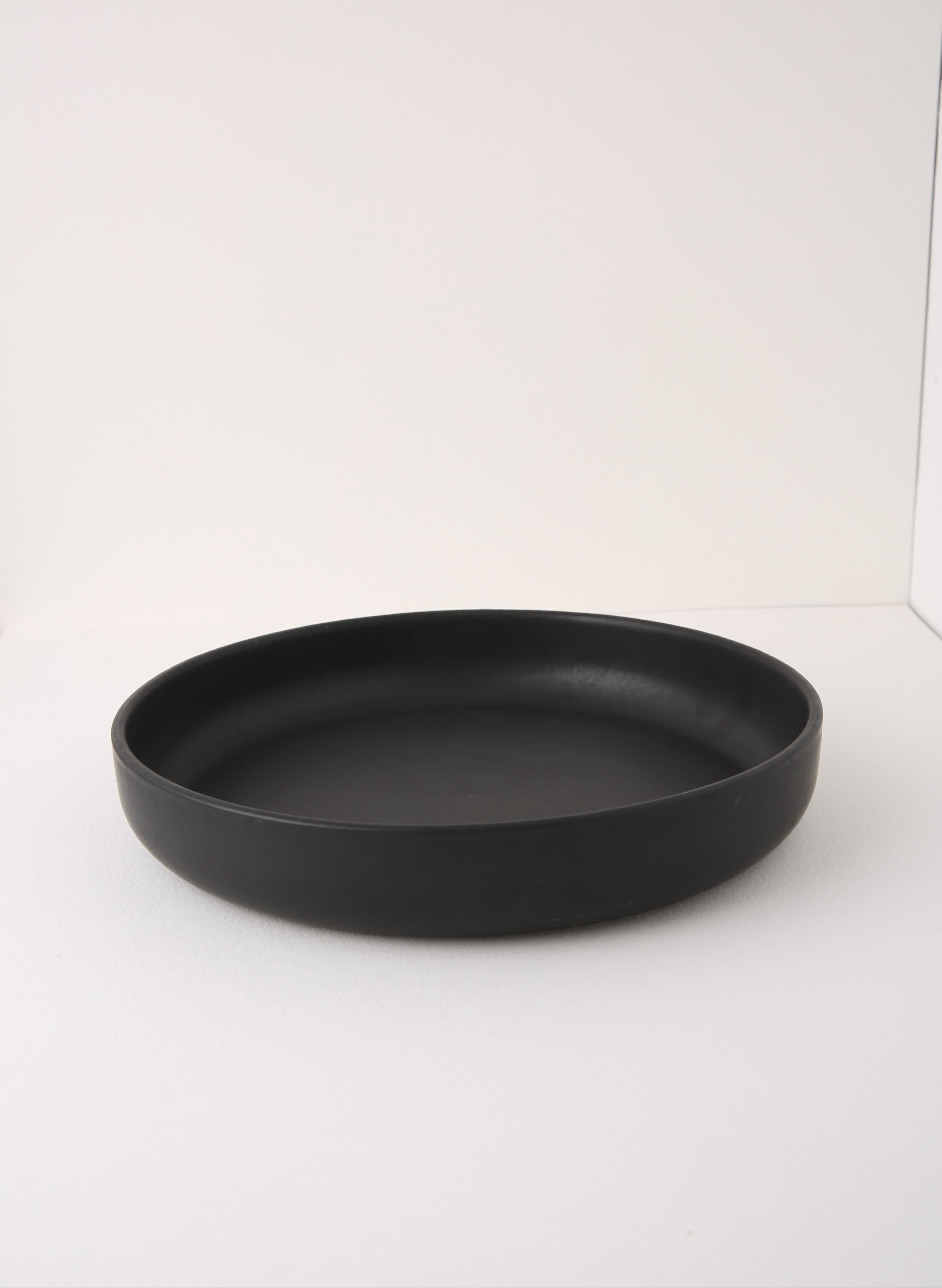 A matte black shallow round tray or plate, photographed from a side angle on a plain white studio background.