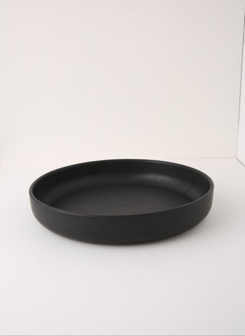 A matte black shallow round tray or plate, photographed from a side angle on a plain white studio background.
