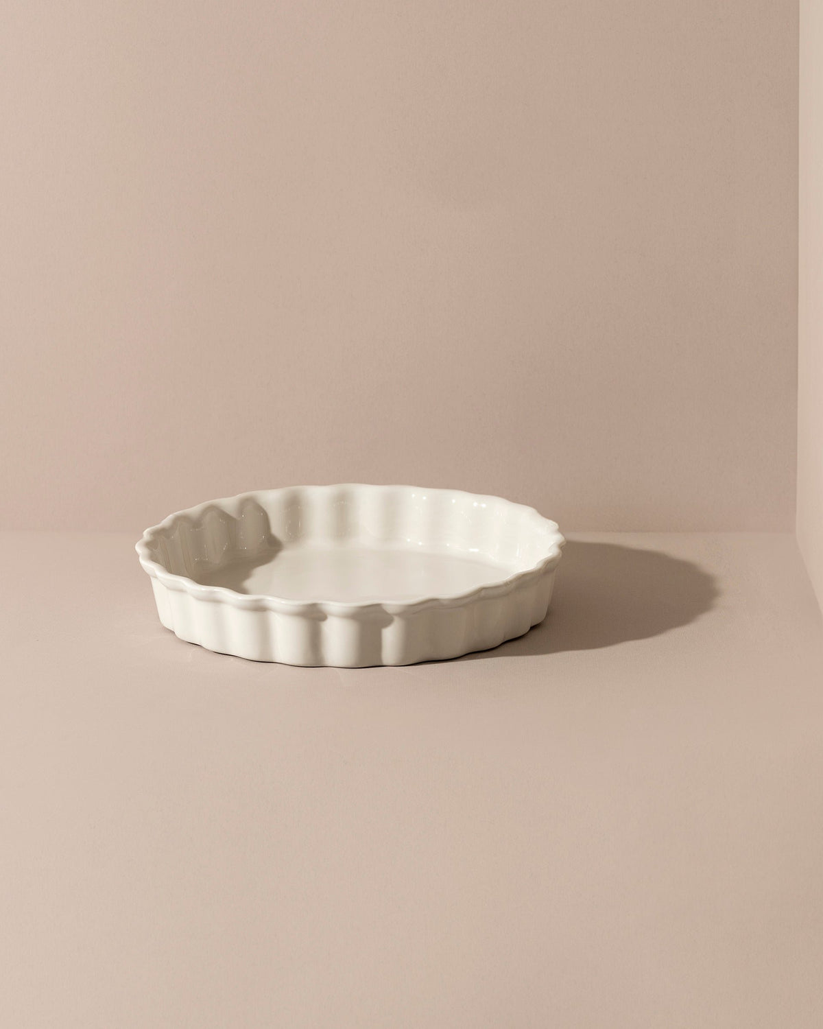A white rounded stoneware pie dish with wavy side walls displayed on a neutral background, accompanied by a wooden cutting board with a pie on it, some garlic bulbs, and a small white bowl.