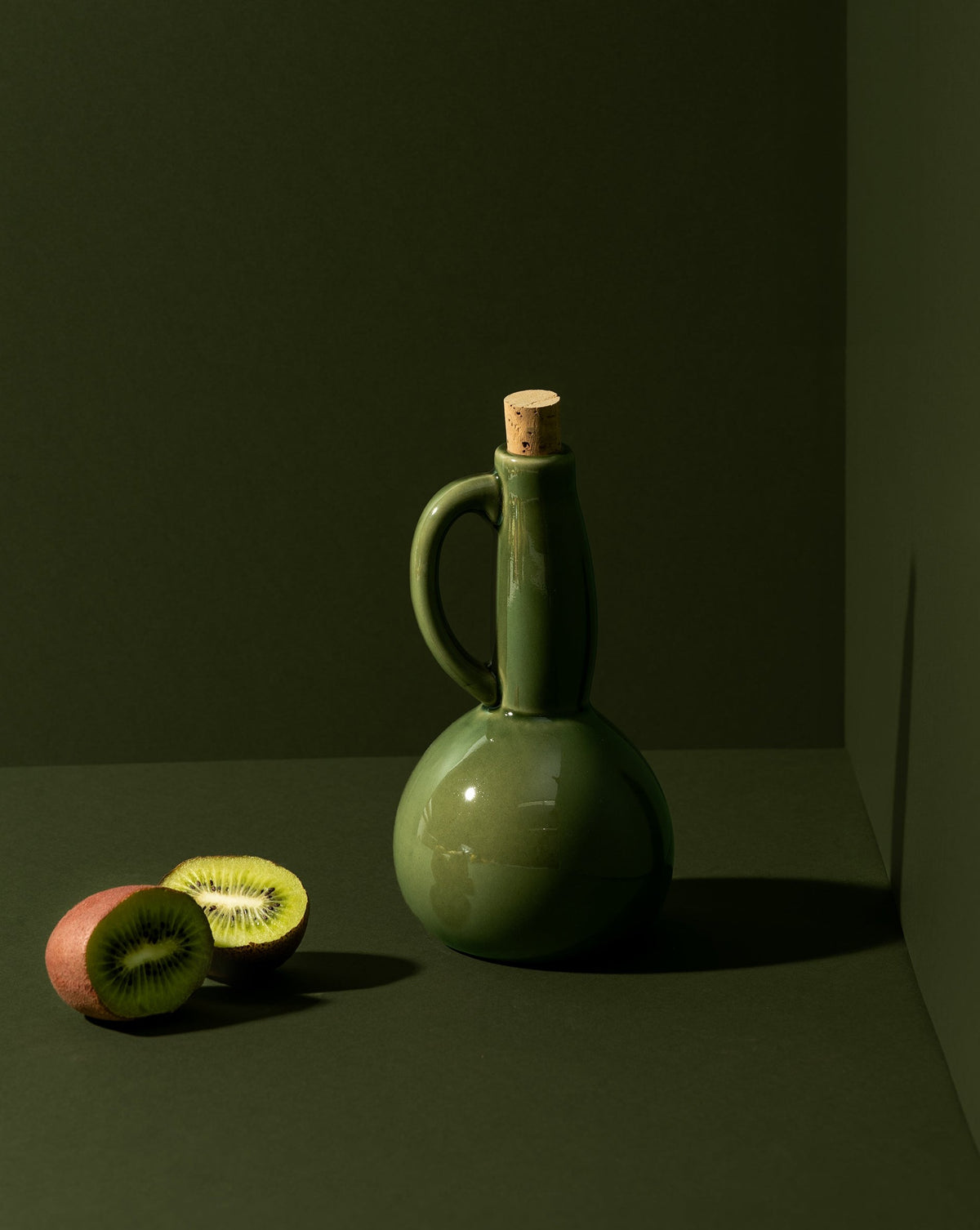 A white ceramic olive oil dispenser with a matte finish, a cork stopper, and a handle, displayed in a corner setting with nuts scattered in front.